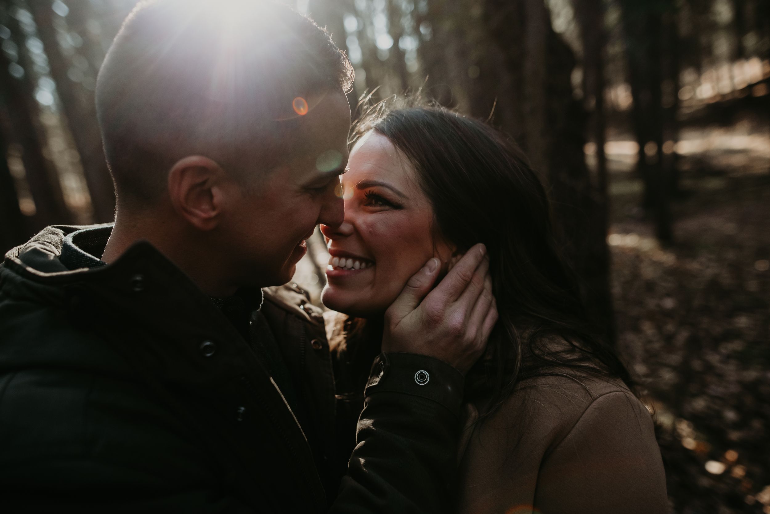 A laughing couple shares a tender moment in sunlit woods during an outdoor engagement photo session.