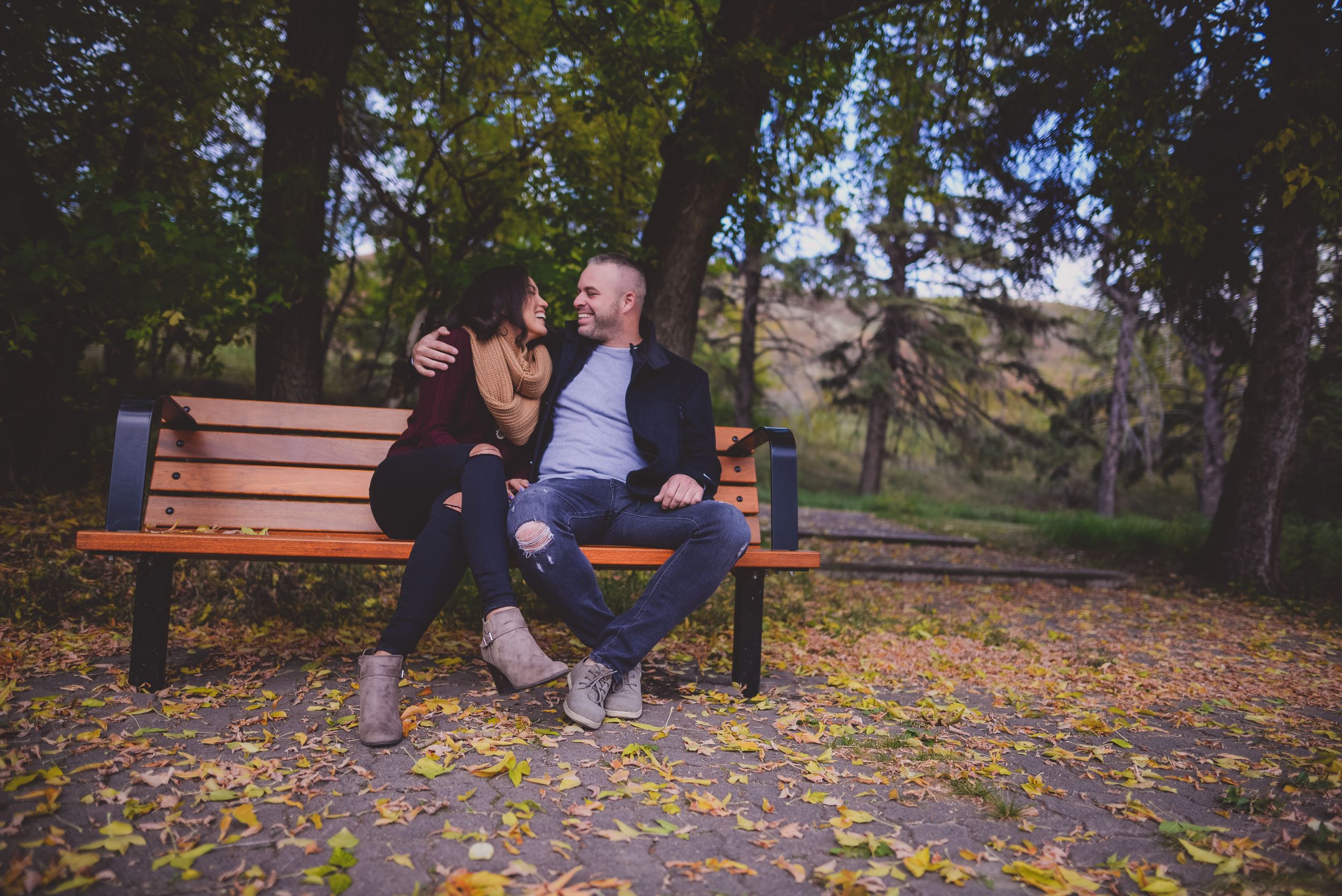 Happy couple sitting together on a wooden park bench surrounded by fallen autumn leaves and trees.