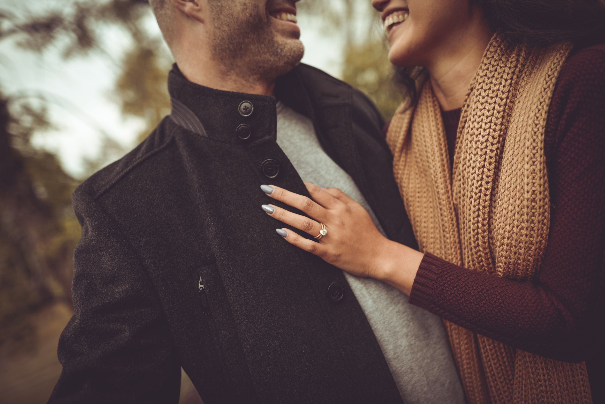 Close-up of smiling couple embracing outdoors, woman wearing engagement ring resting hand on man's chest.