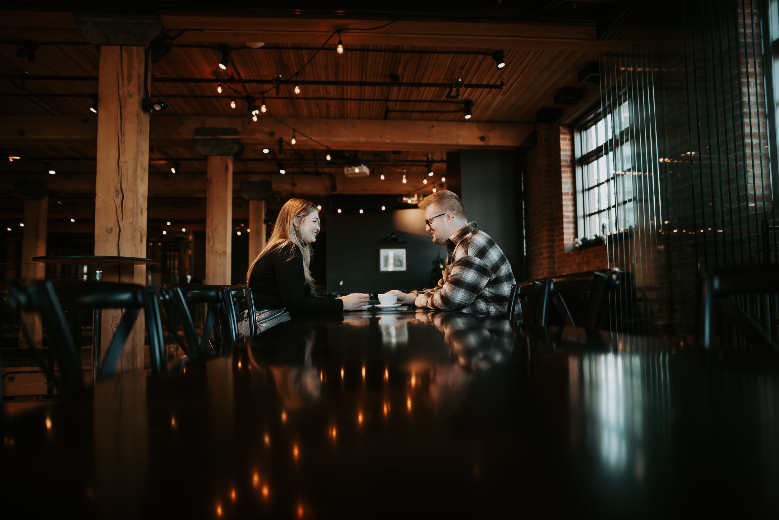 A couple shares a romantic moment across a dark wooden table in a warmly lit industrial venue.