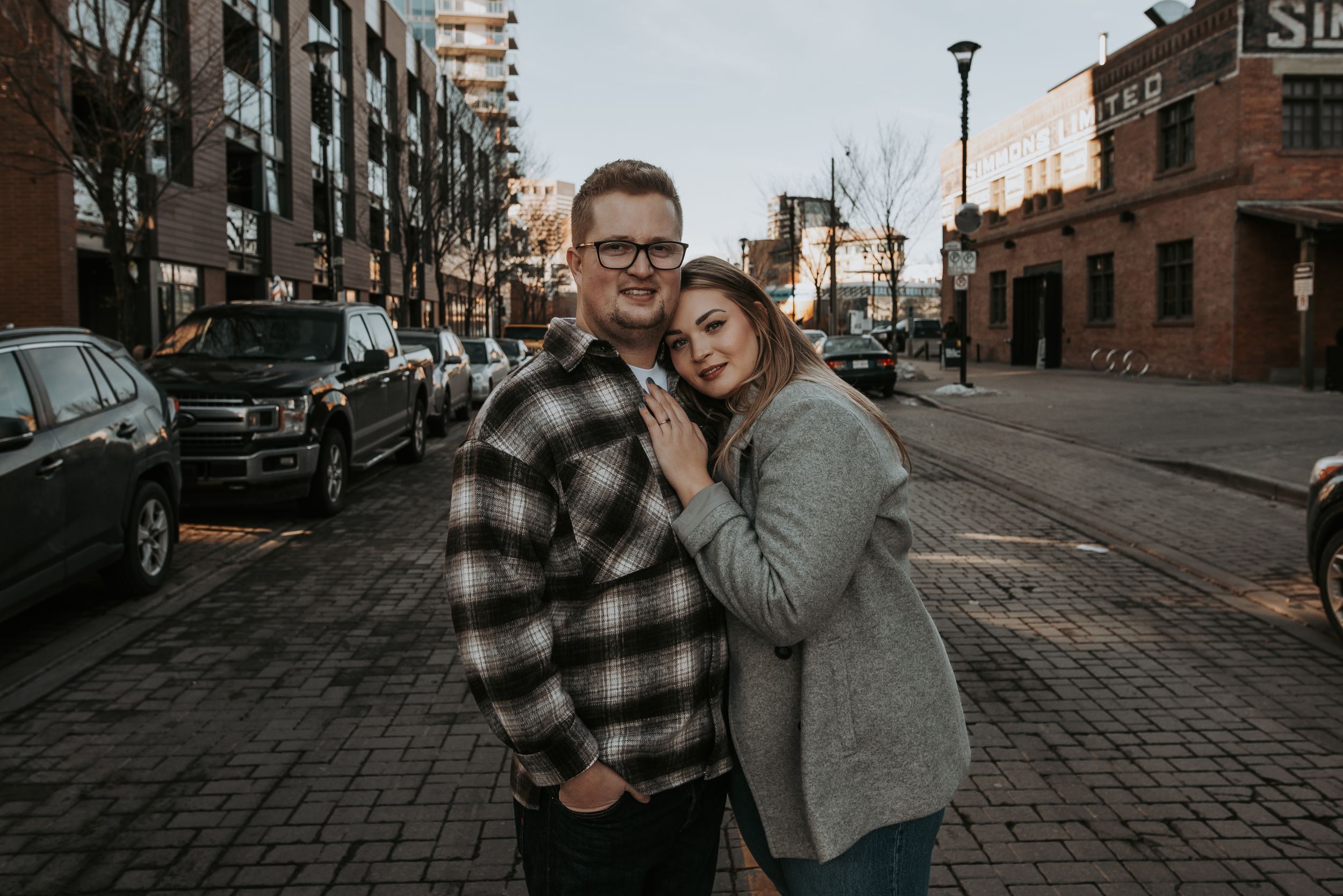 A couple poses on a cobblestone street in an urban setting with city buildings in the background.
