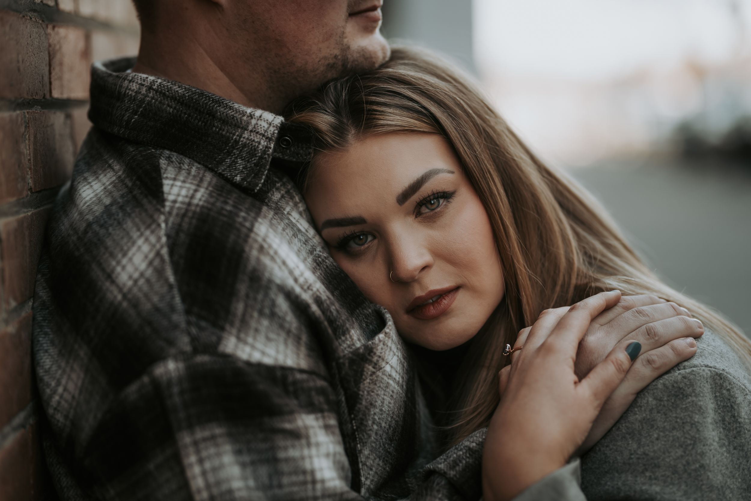 A woman leans against a man in a plaid shirt by a brick wall in an intimate couple portrait.