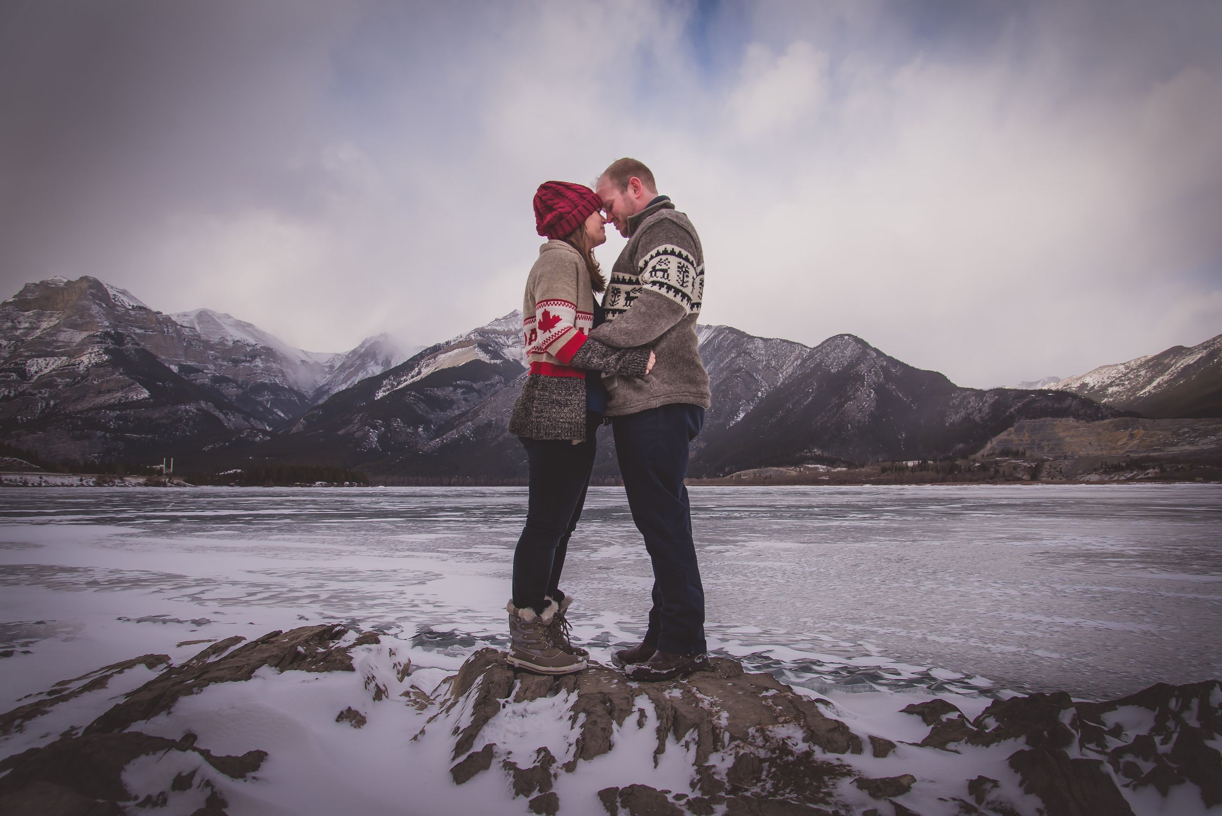 Couple sharing romantic kiss on frozen lake with snowy mountains in background.