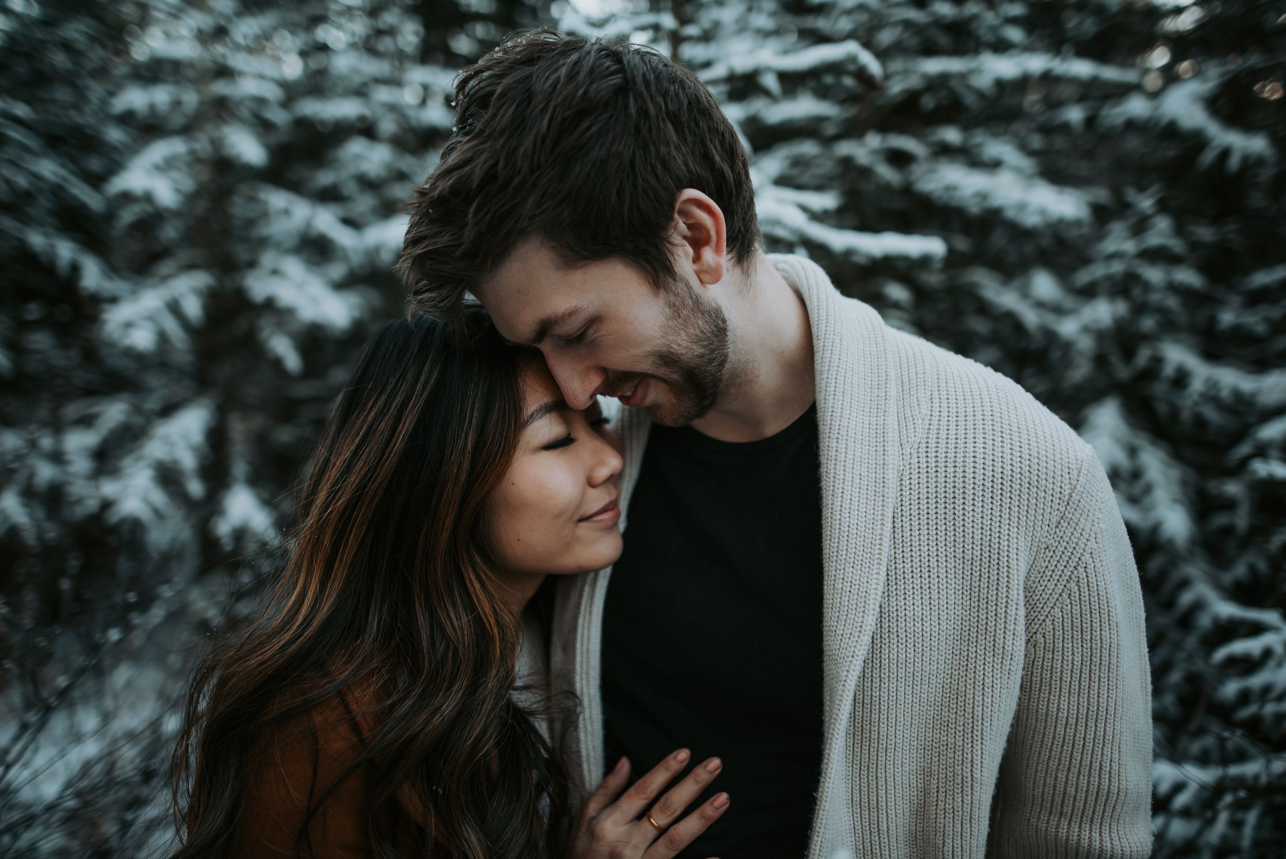 A couple shares a romantic forehead touch surrounded by snow-covered trees in a winter portrait.