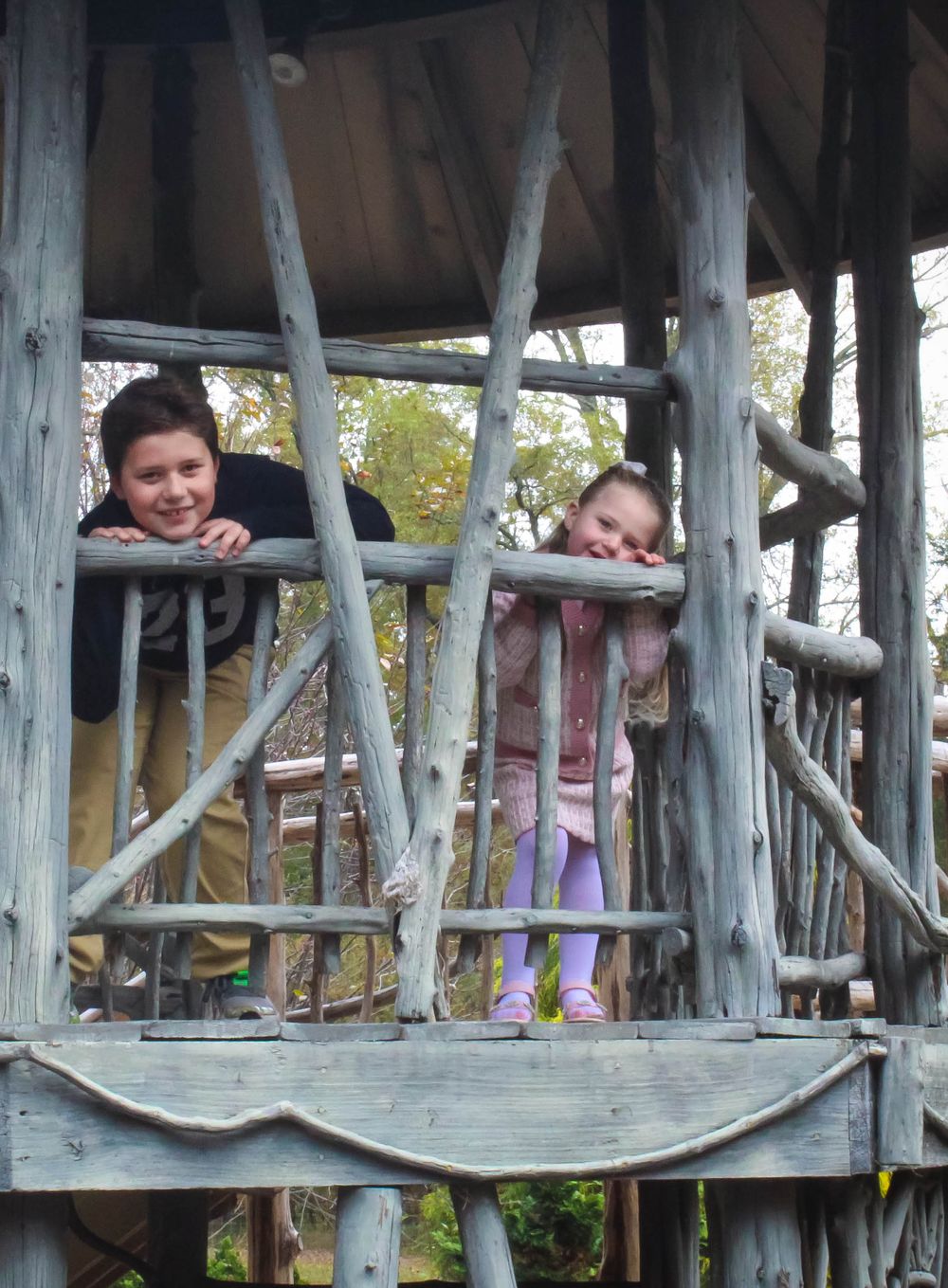 Two young people lean over wooden railings of a rustic treehouse structure.