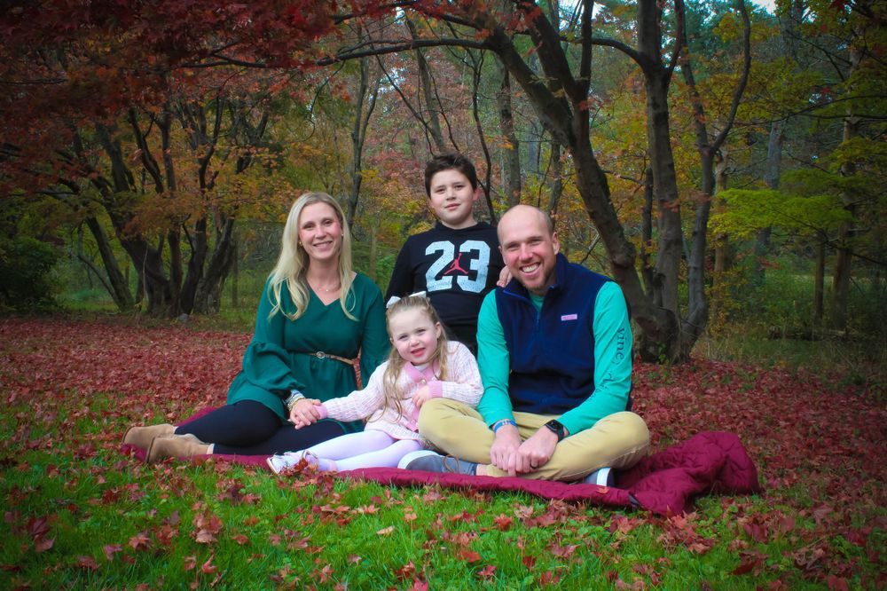 Family of four poses together on ground with colorful fall foliage in background.