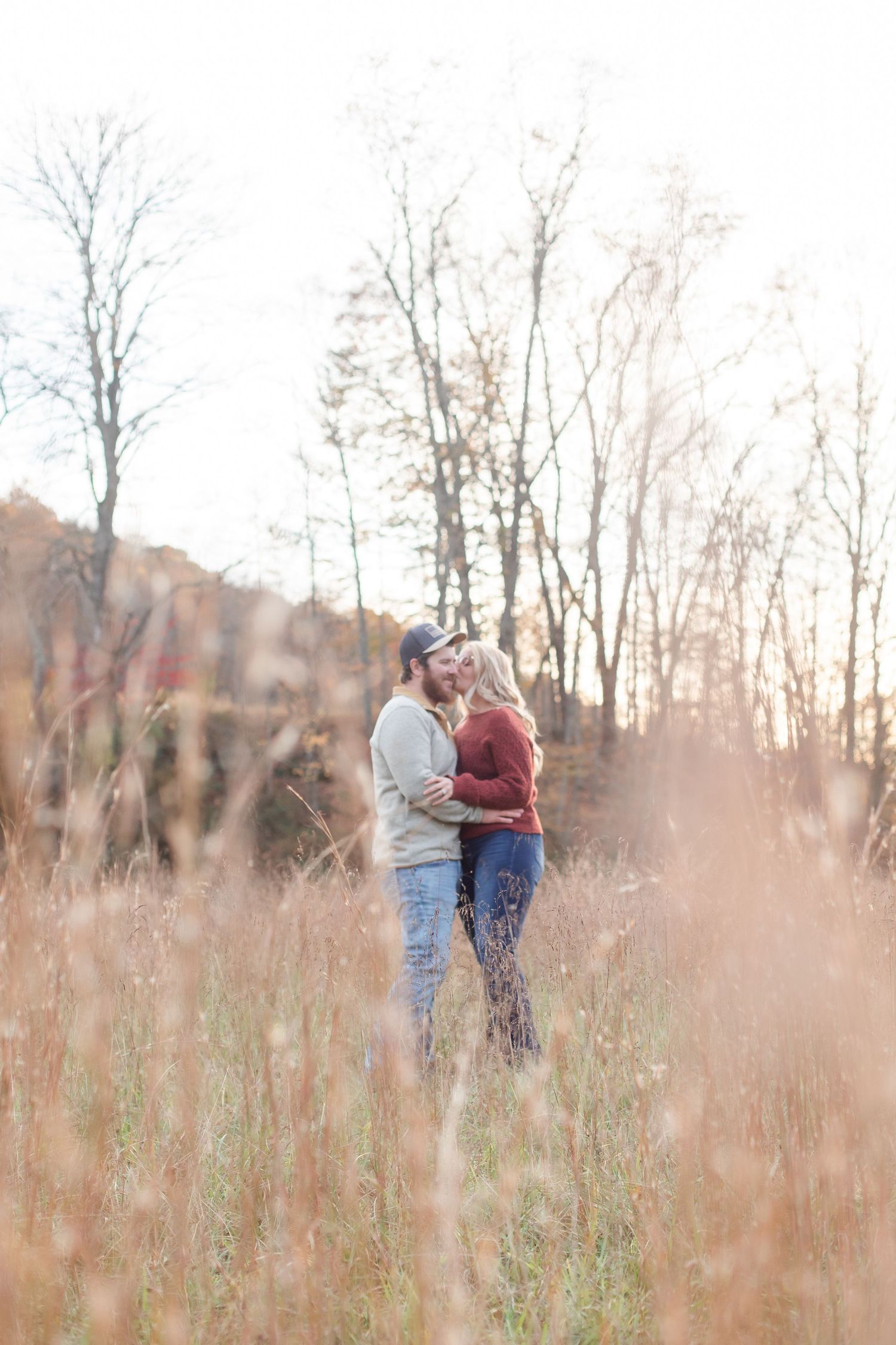 A romantic couple poses in a field of tall grass during golden hour with autumn trees in the background.