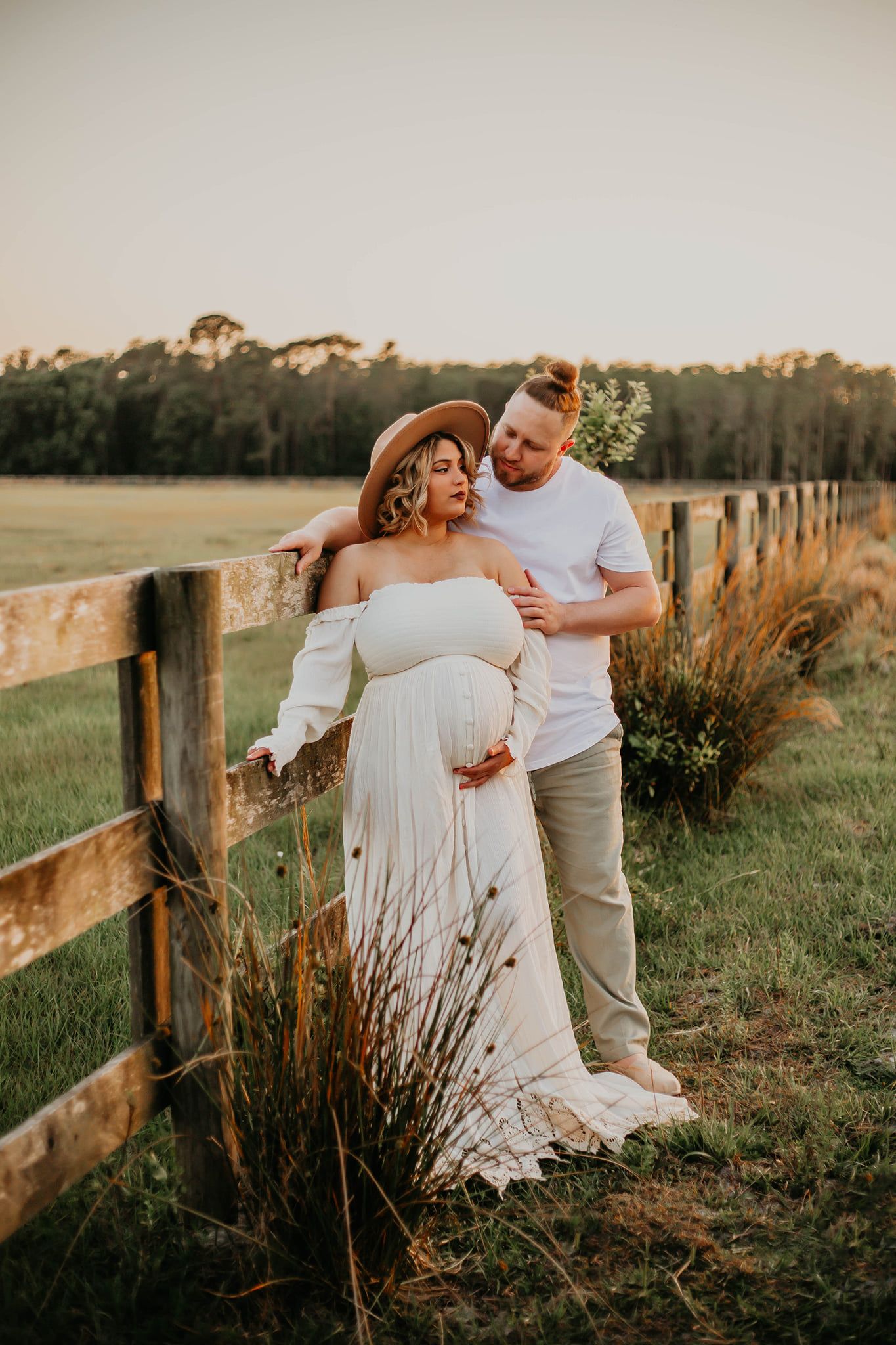 Rustic maternity photo of couple by wooden fence during golden sunset in countryside setting.