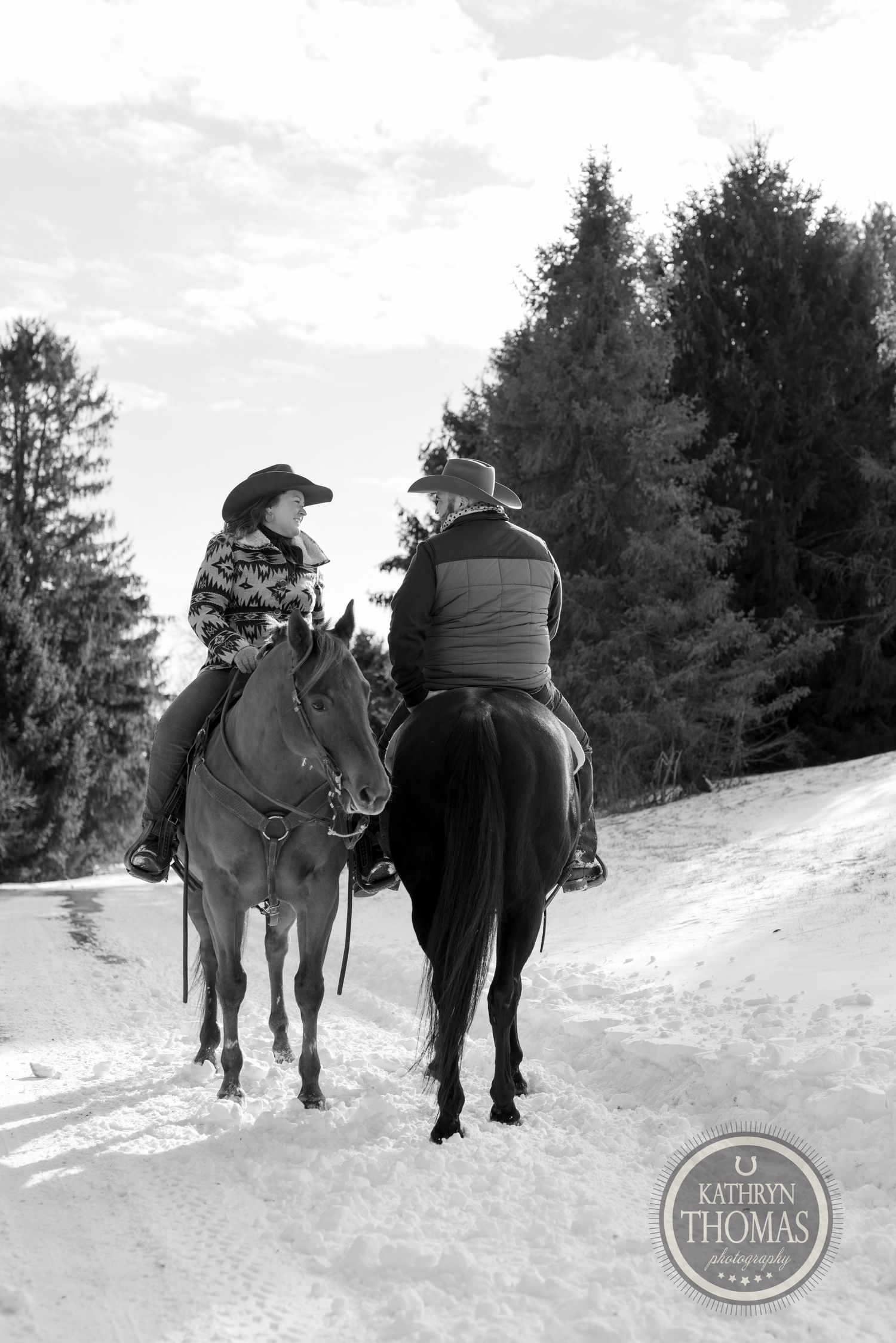 Two riders on horseback chat while riding along a snowy forest path in winter.