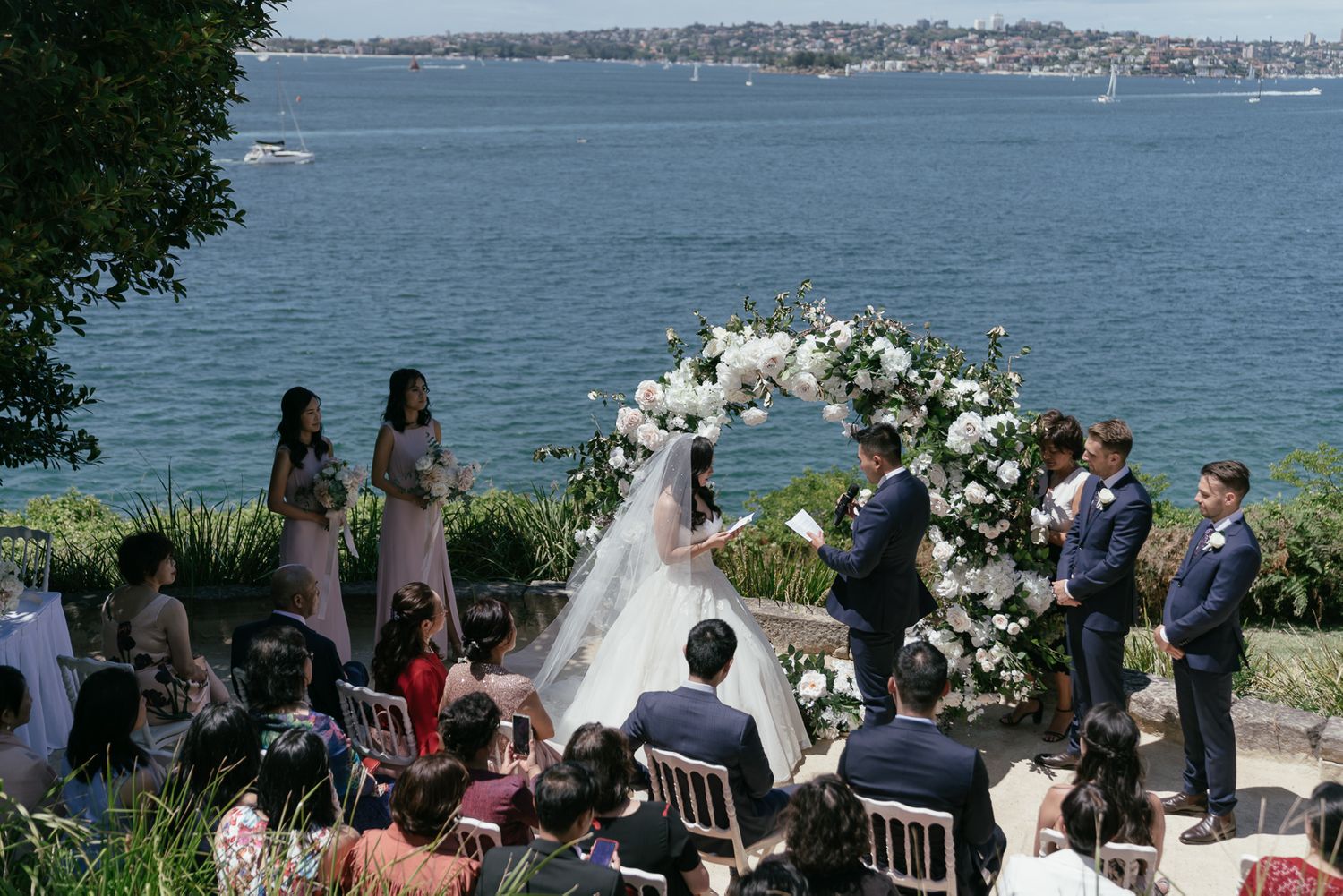Outdoor waterfront wedding ceremony with white floral arch and Sydney Harbour views.