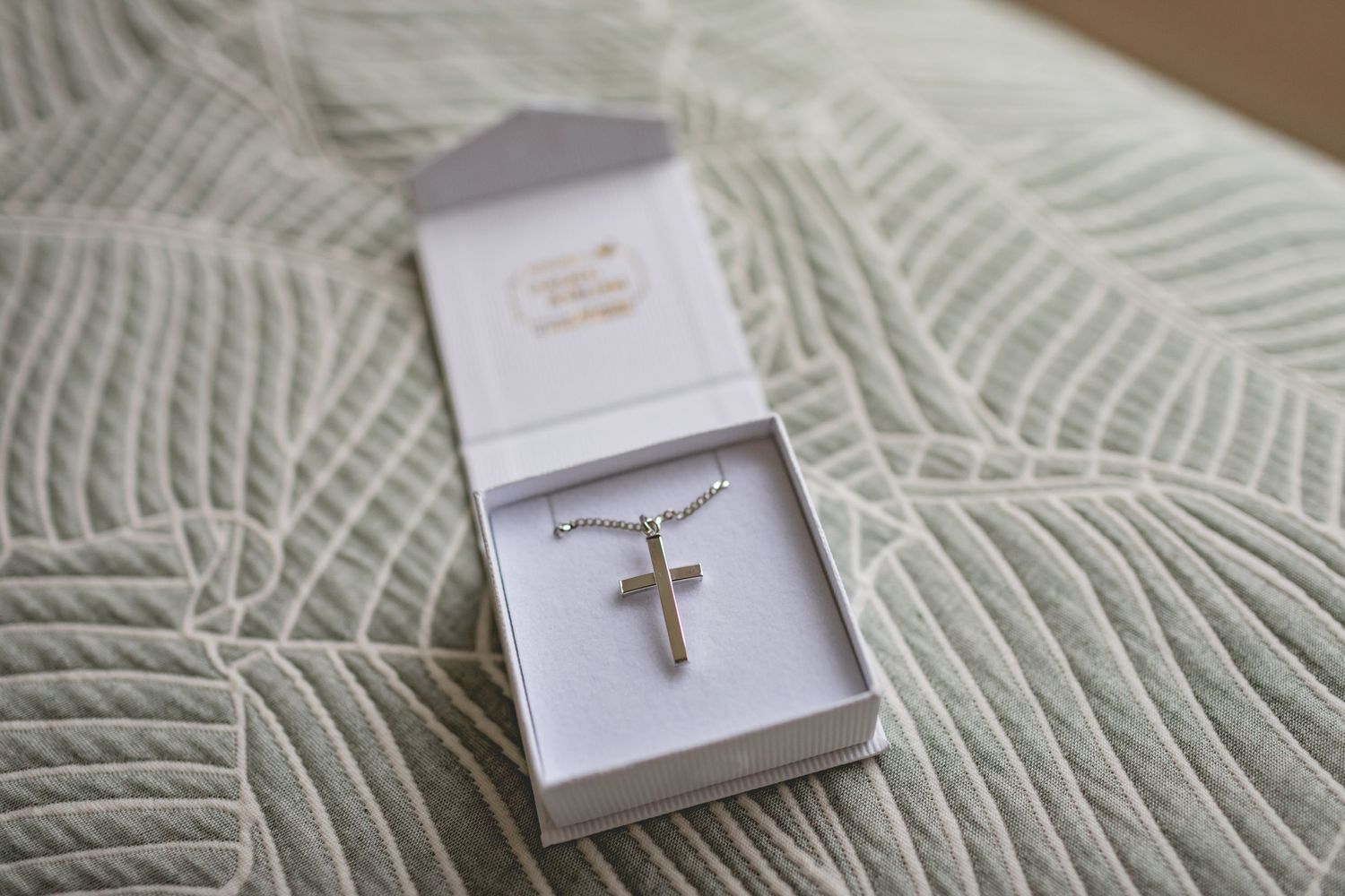 A silver cross necklace pendant displayed in a white gift box on a textured knit blanket.