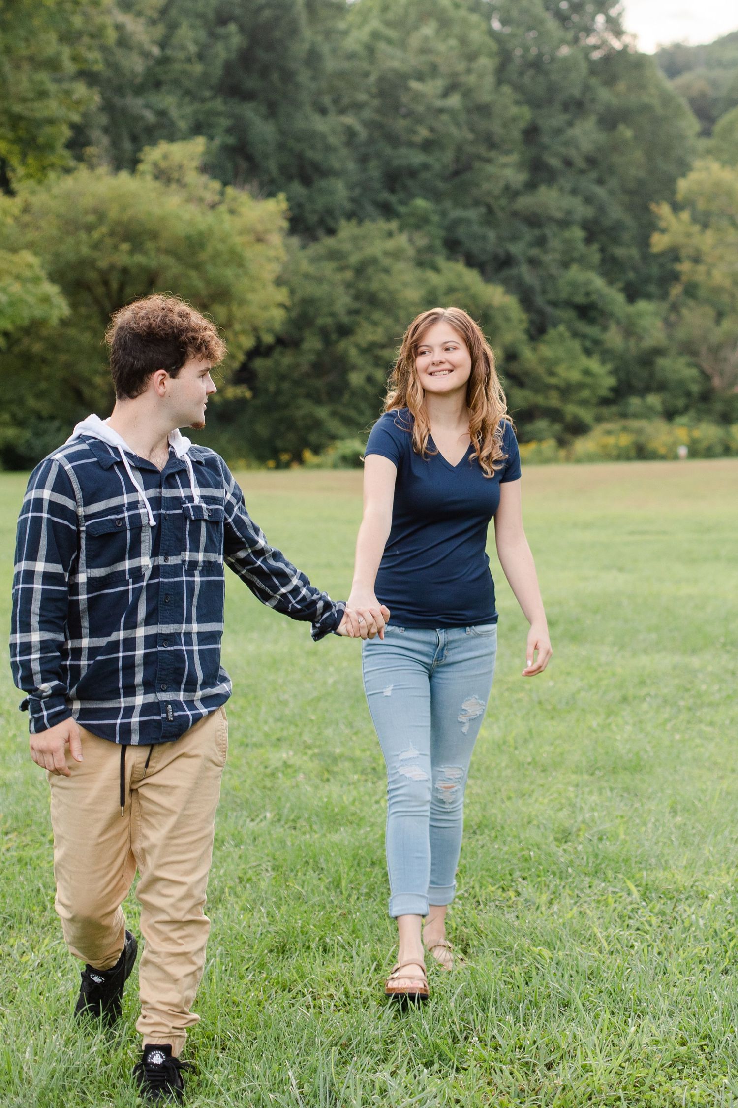Playful couple walking and laughing together in a grassy field surrounded by trees.