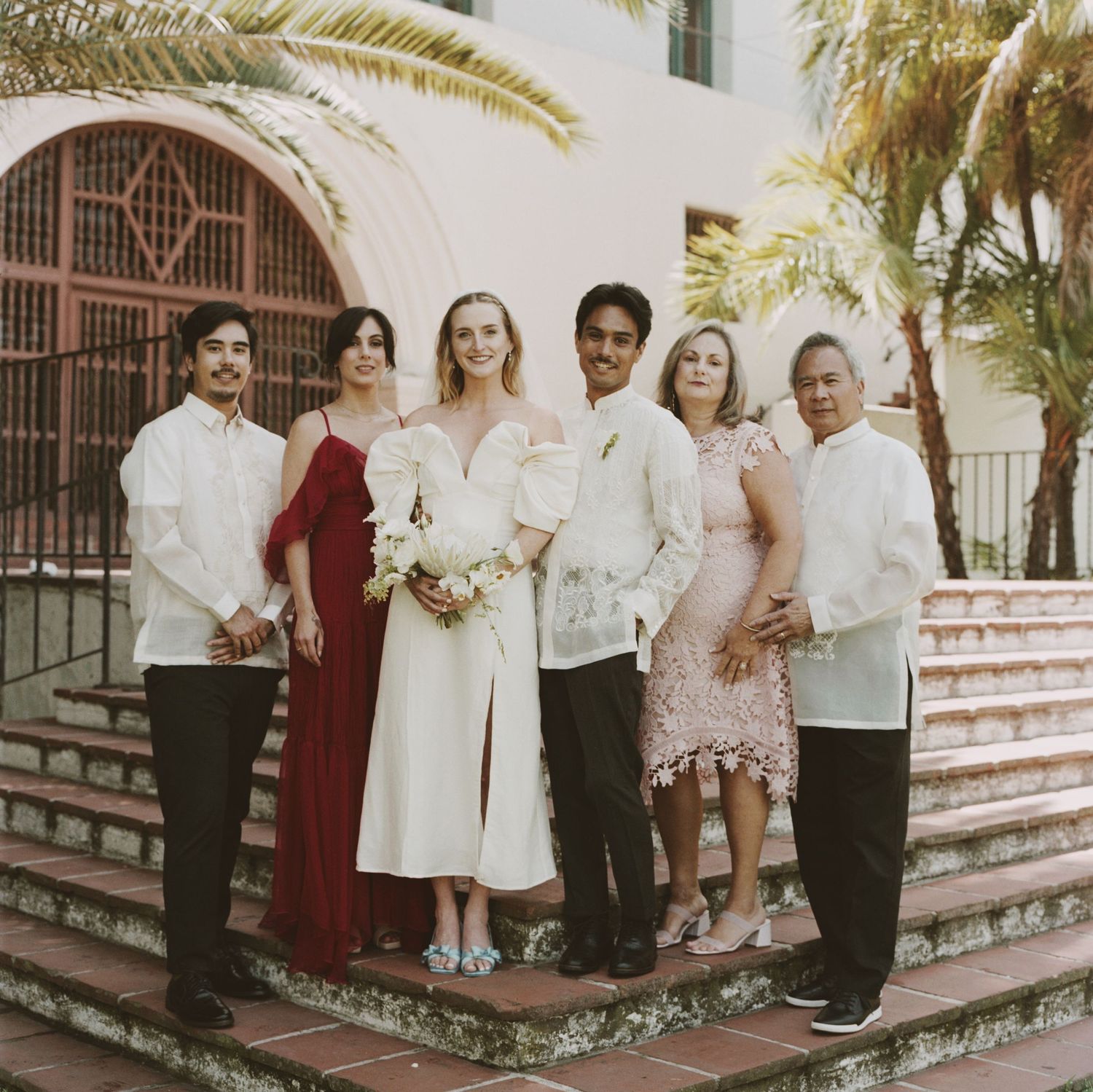 Wedding party in formal attire poses on steps of Spanish colonial style building with palm trees.