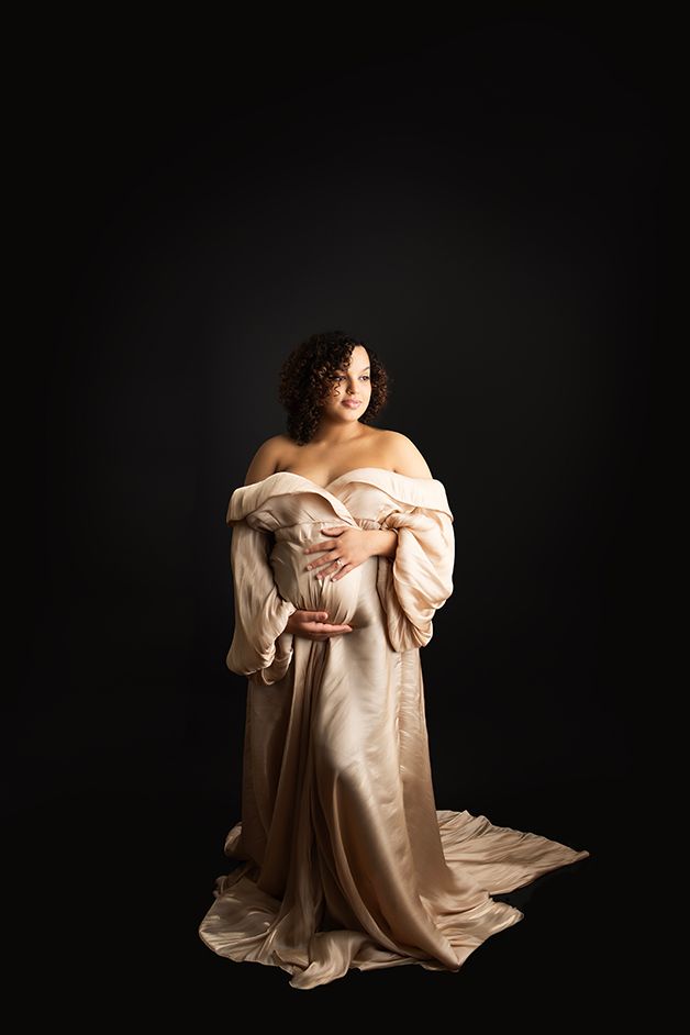 Image of a pregnant mother standing on a black backdrop wearing a champagne colored maternity gown.
