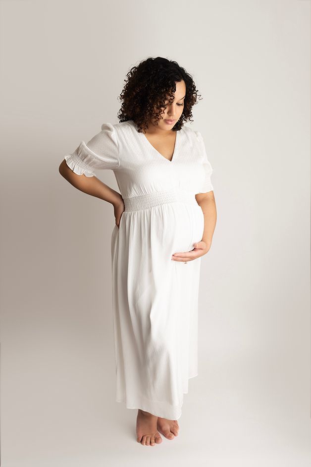Pregnant mom standing on a white backdrop wearing a white dress.  Holding her belly and looking at her belly.