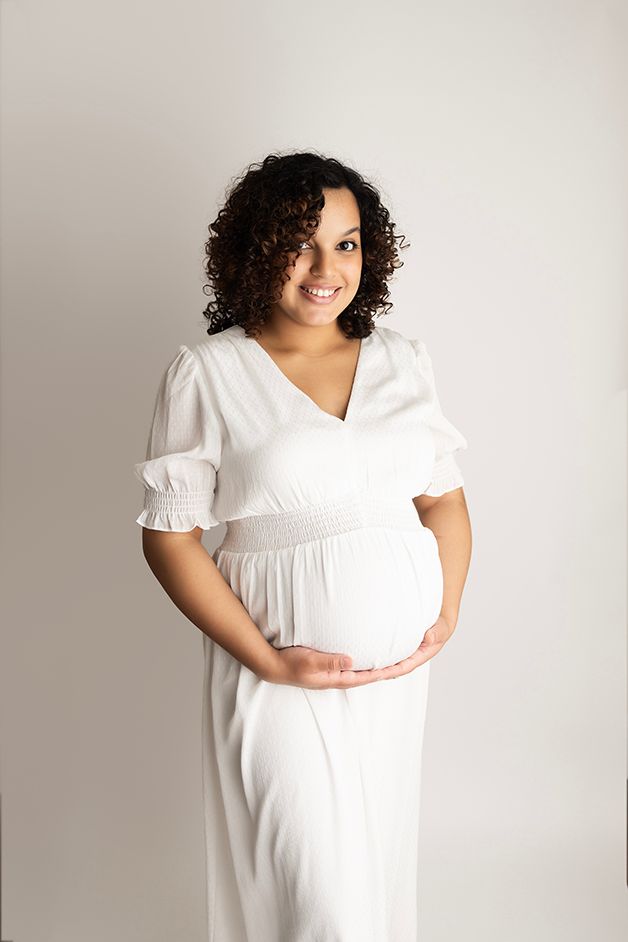 Pregnant mom on a white backdrop in a white dress holding her belly and smiling at the camera. '