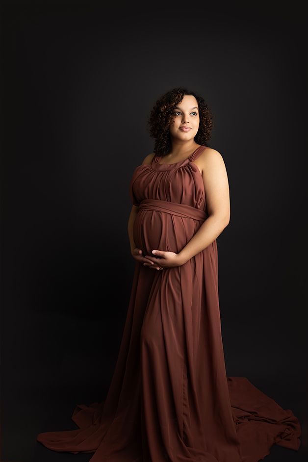 Image of pregnant mother craddling her belly.  She is standing on a black backdrop in a maroon sleeveless dress.