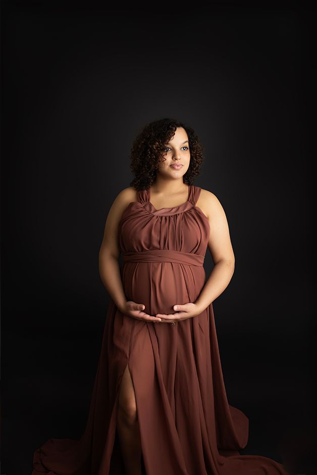 Pregnant mom in a maroon dress standing, holding her belly on a black background.