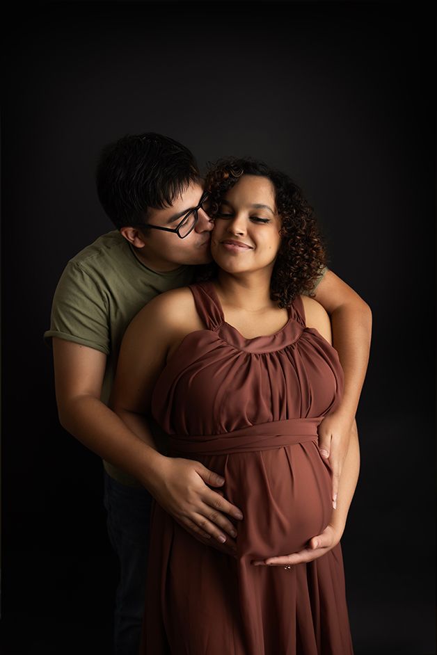Pregnant mom being held by dad.  Dad is kissing mom's cheek.