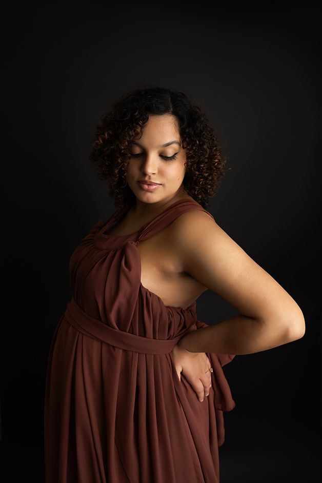 Pregnant mom standing to the side showing her belly in a maroon dress.  Light is cascading down her face.