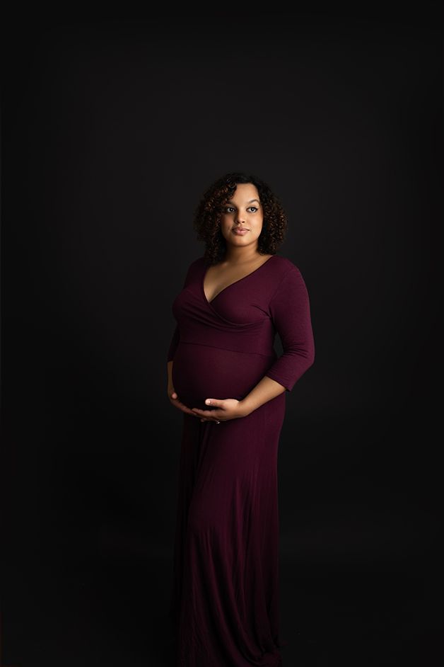 Pregnant Mom standing on a black backdrop in a burgundy dress, majestically looking towards the light.