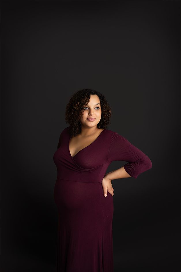 Pregnant mom standing on a black backdrop, hand resting on her hip, showing her belly.