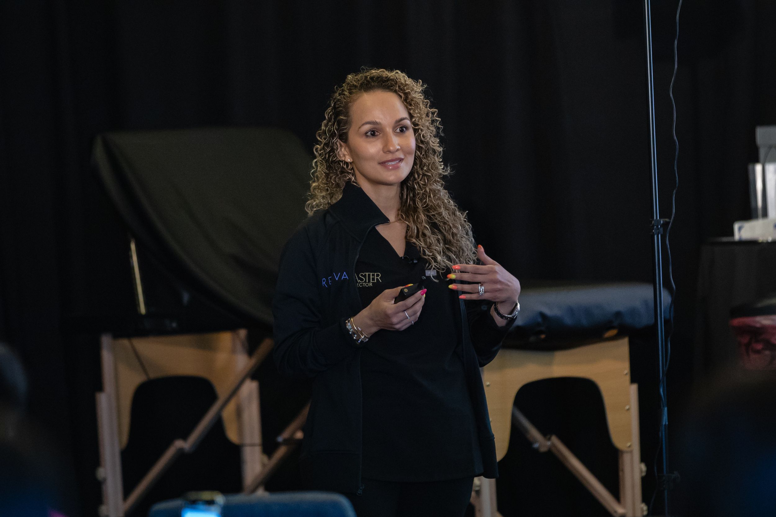 A speaker in a black sweater gestures while presenting on stage with a black backdrop.