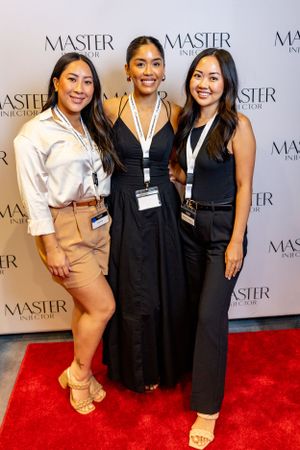 Three women pose together on red carpet at Master event wearing dressy casual outfits and name badges.