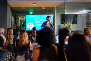 Crowd watches presenter on stage with turquoise backdrop at corporate event.