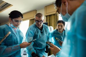 Medical students in blue scrubs gather around looking at their mobile phones during a training session.