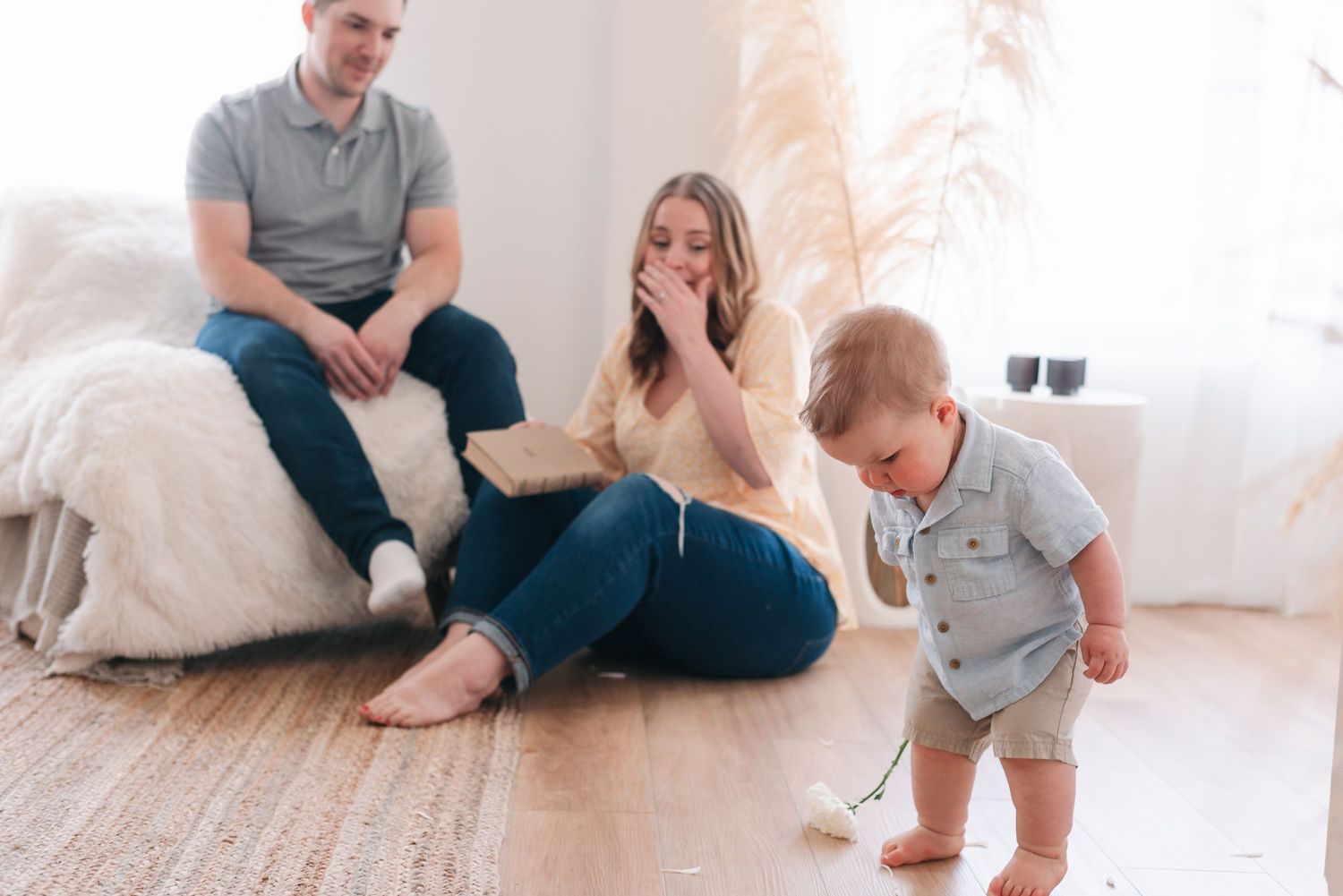 Toddler taking first steps in bright living room as parents watch from cushions on floor.
