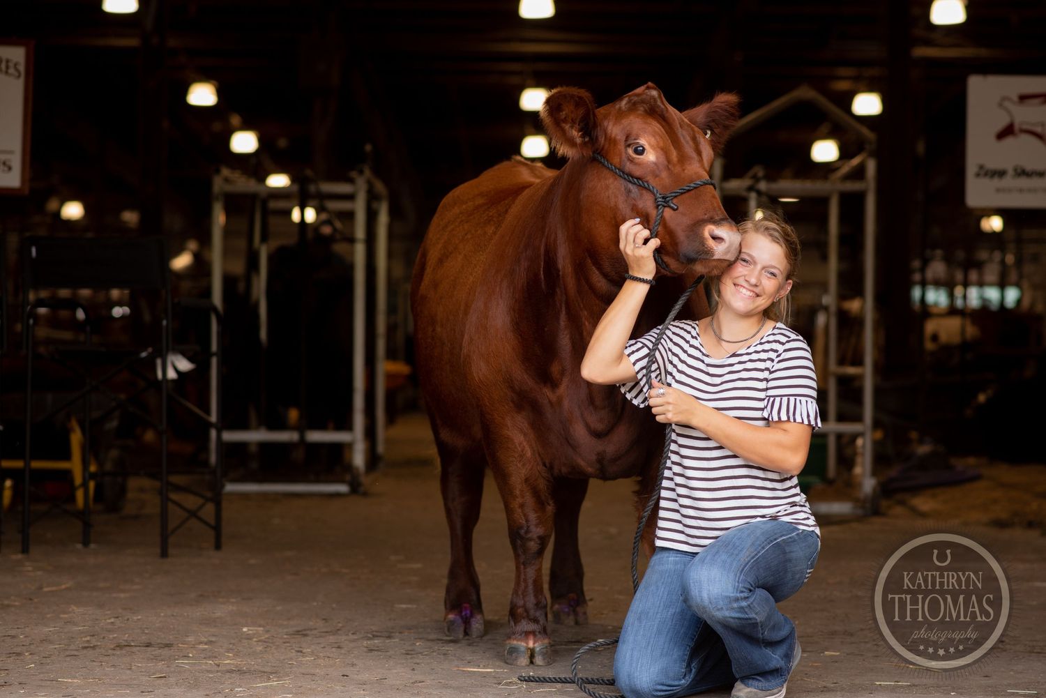 A person in a striped shirt poses with their show cow in a barn setting.