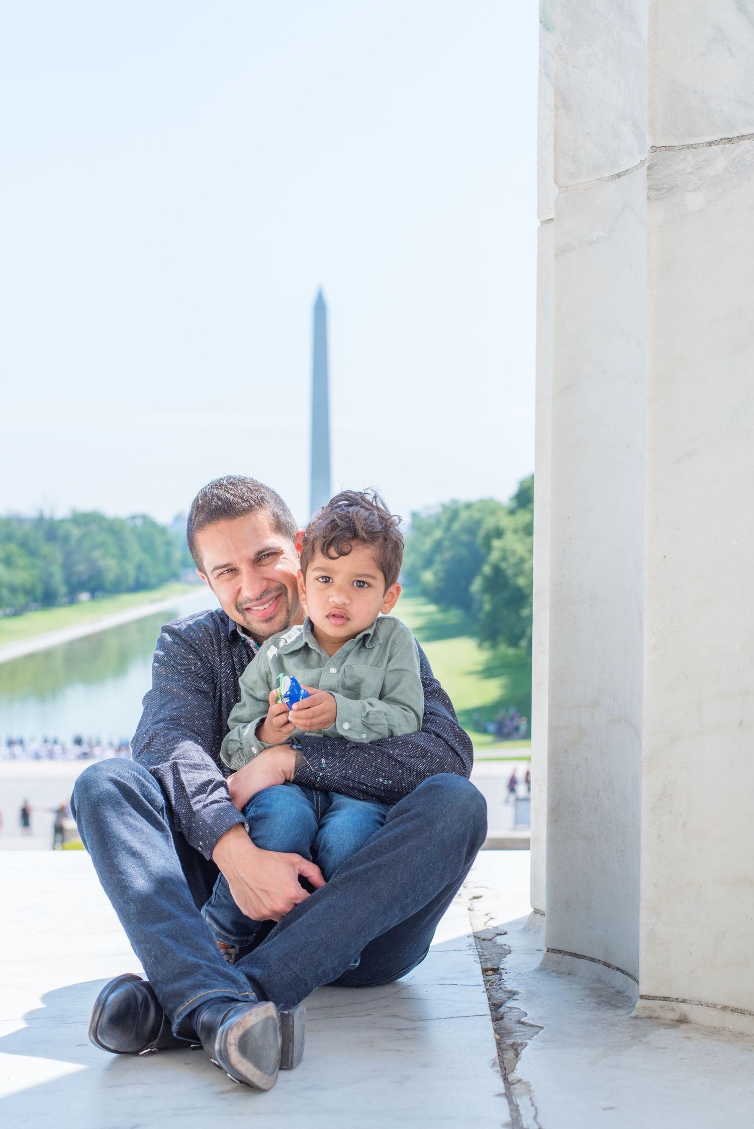 on ground, dad holds son in his lap, with Washington Monument behind them. Son holds a toy car
