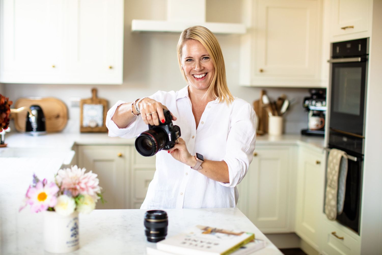 A content creator holds a professional camera while working in a bright modern kitchen with flowers on the counter.