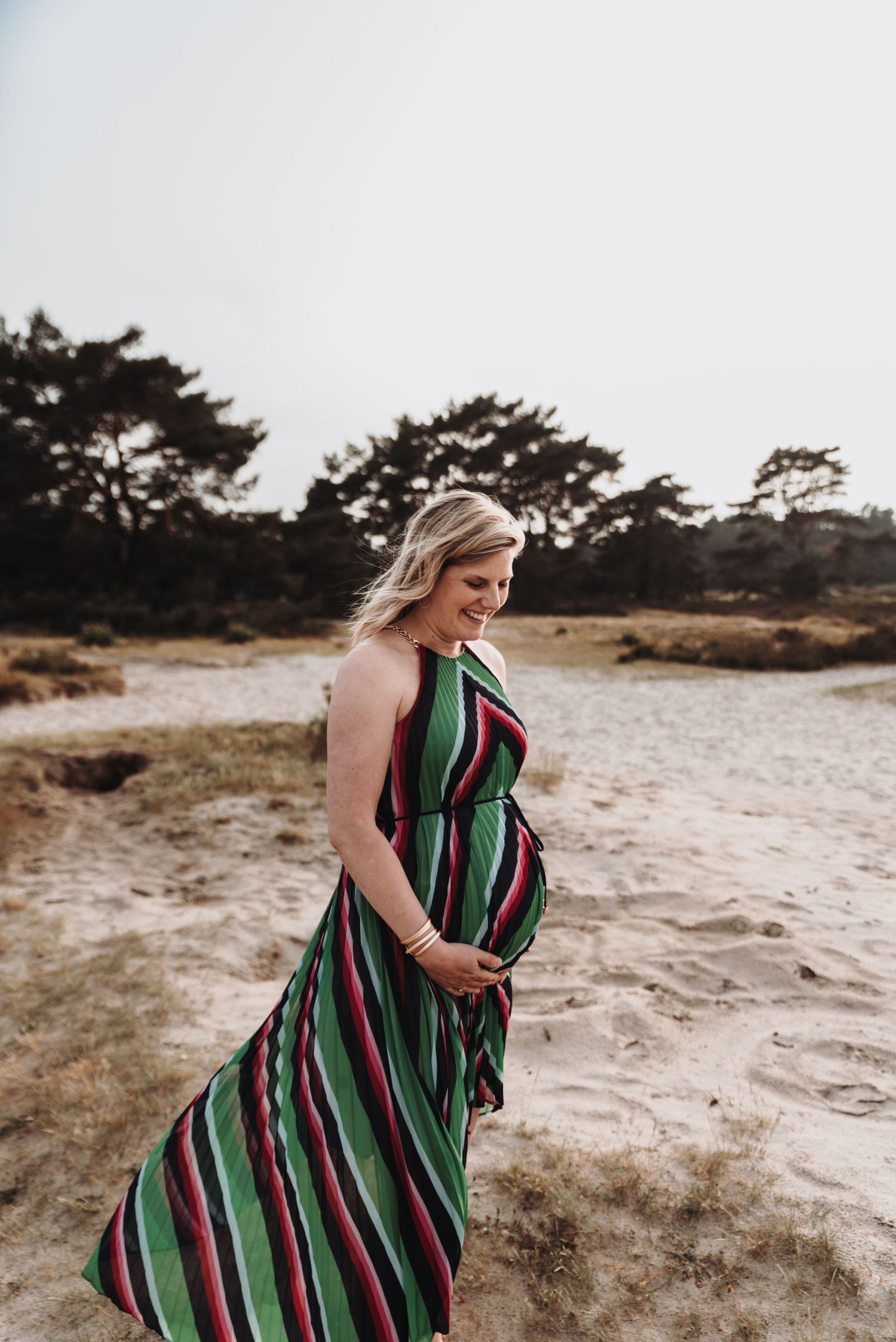 Expecting mother in striped maxi dress poses on sandy beach at sunset.