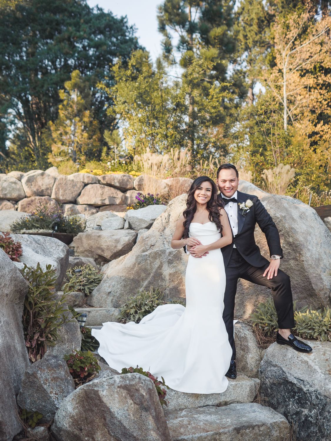 A newlywed couple poses on rocky landscaping surrounded by autumn trees and natural scenery.