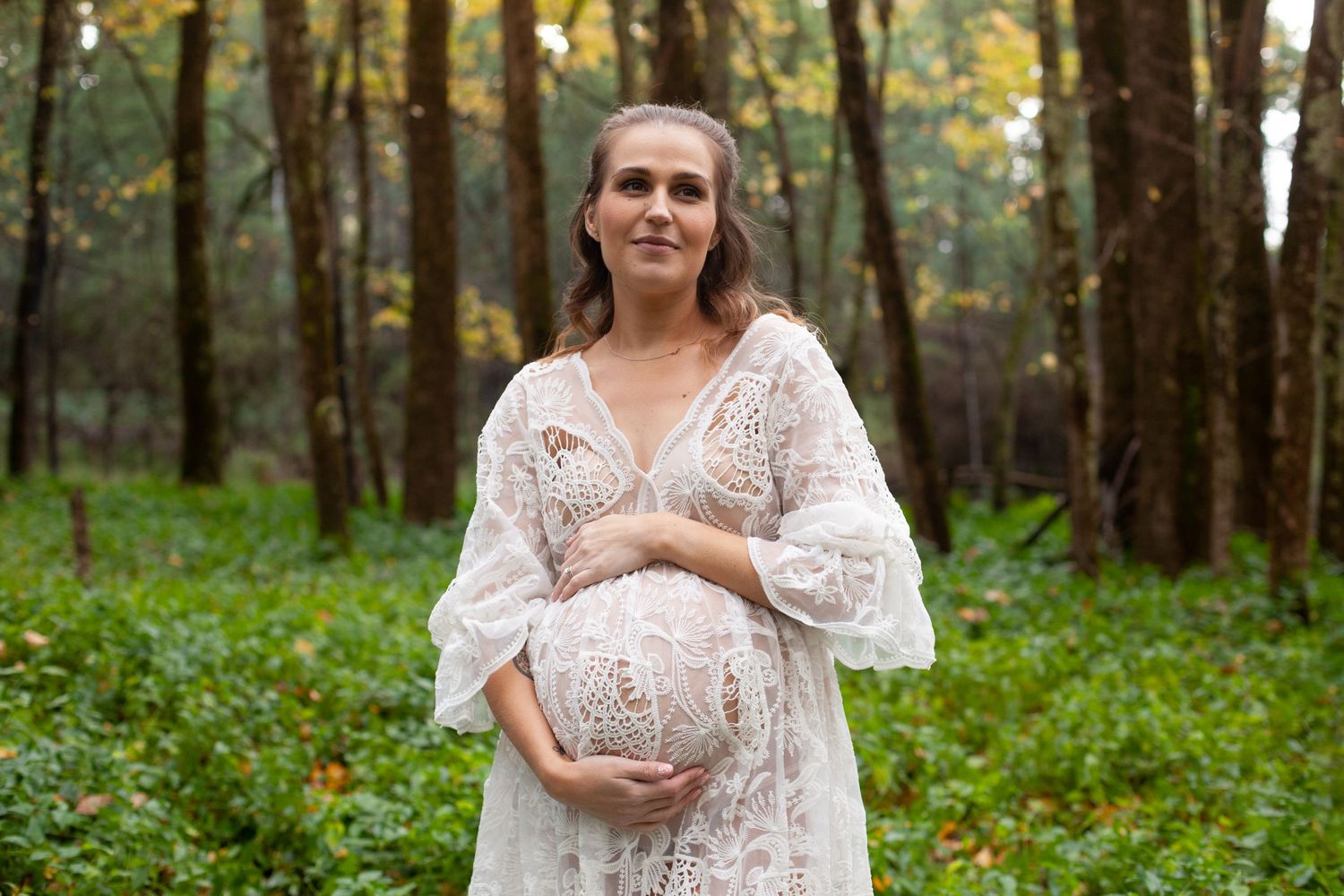 Expectant mother in a flowing white lace dress poses joyfully in a wooded forest setting.