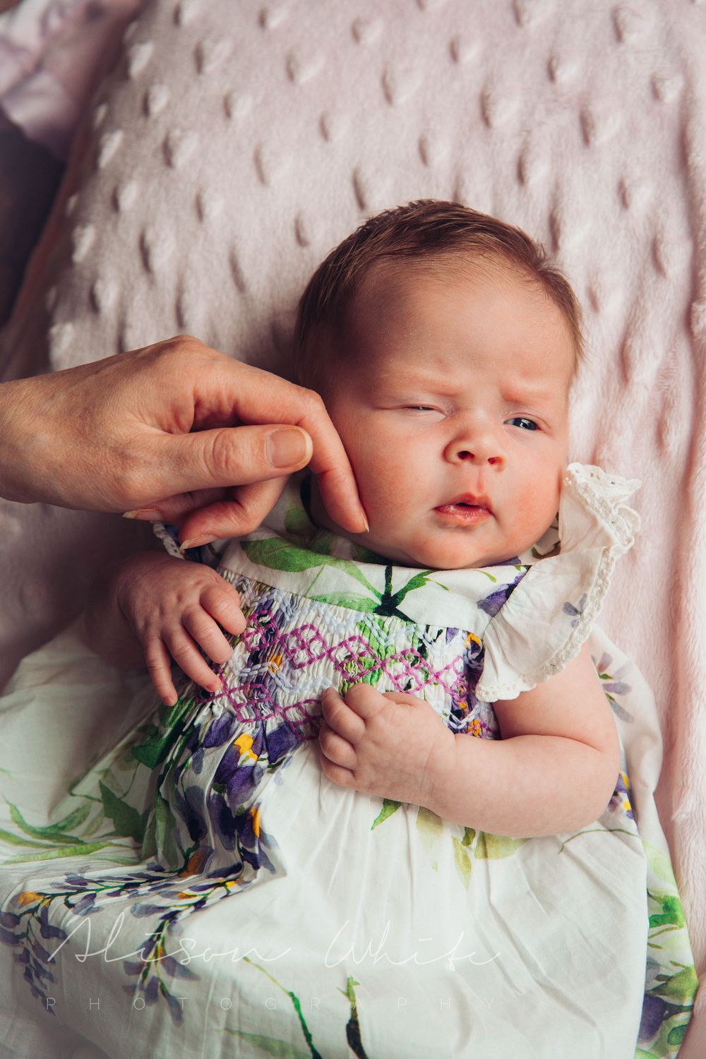 Baby in floral dress lying on pink textured blanket while being touched gently.