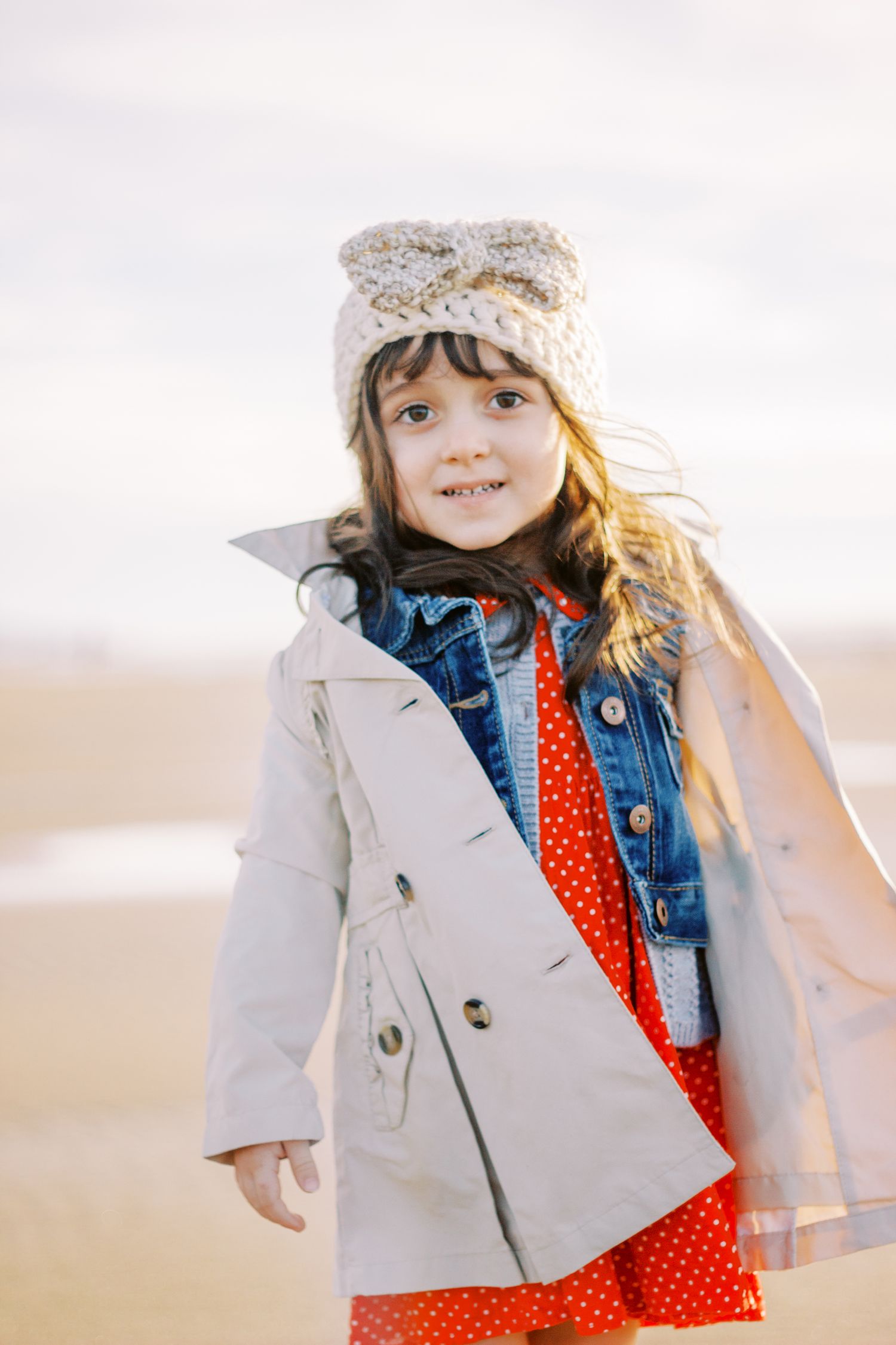 Small child wearing a knit hat, denim jacket and polka dot dress against a light background.