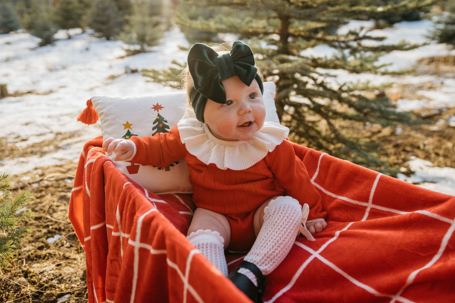 Baby in red dress sitting on blanket outdoors in winter setting.