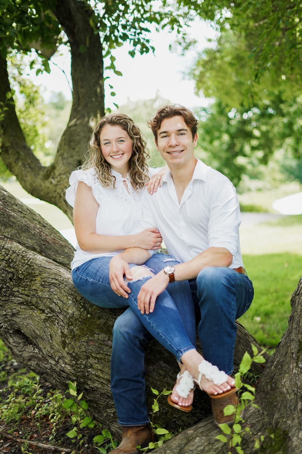 A couple sits together on a large tree branch in a sunny park setting.