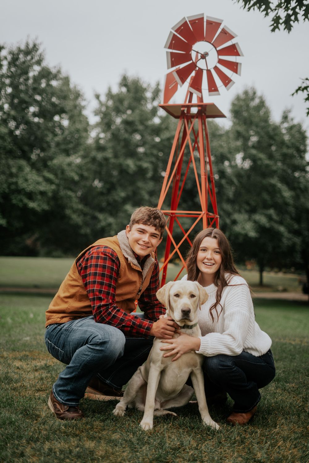 A couple poses with their yellow lab puppy in front of a red windmill.