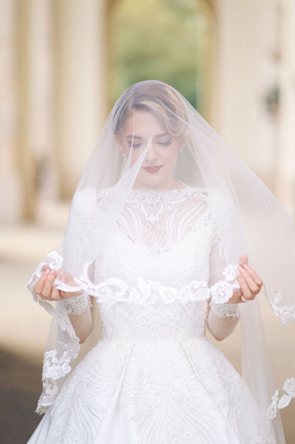 A bride in a white lace gown holds up her delicate veil while standing in an archway.