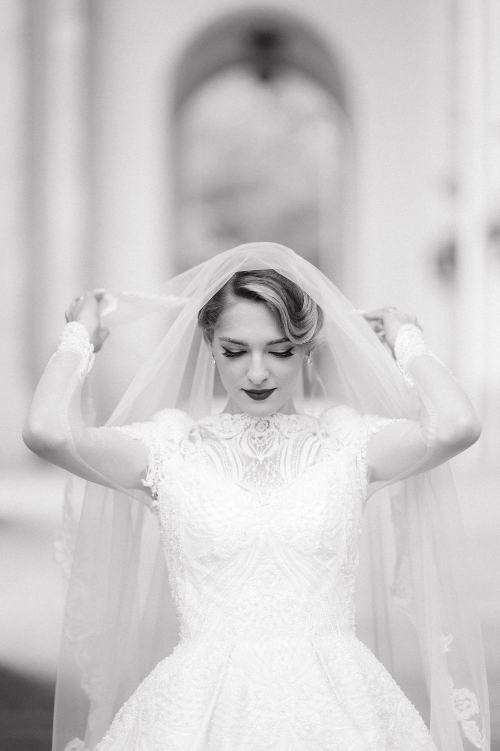 A black and white photograph of a bride adjusting her veil in an elegant pose.