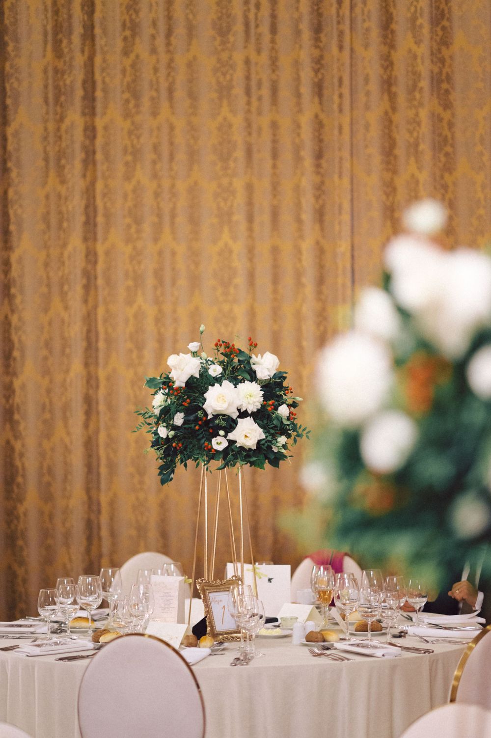 An elegant wedding reception table setting with tall white floral centerpiece and golden drapes.