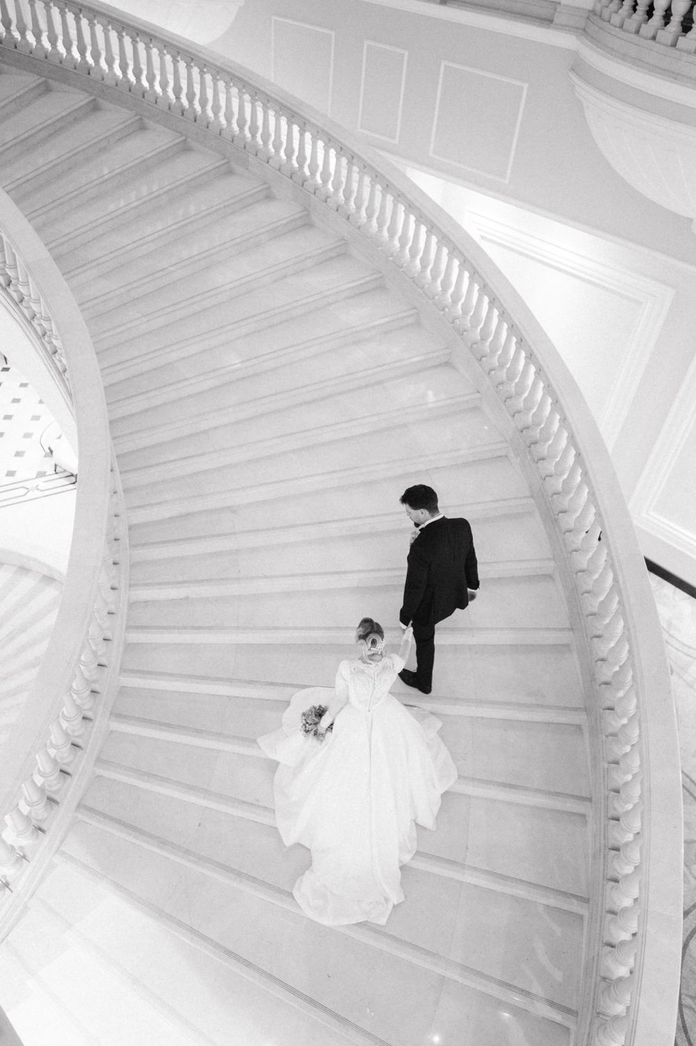 A black and white photo of a couple descending an elegant curved staircase in wedding attire.