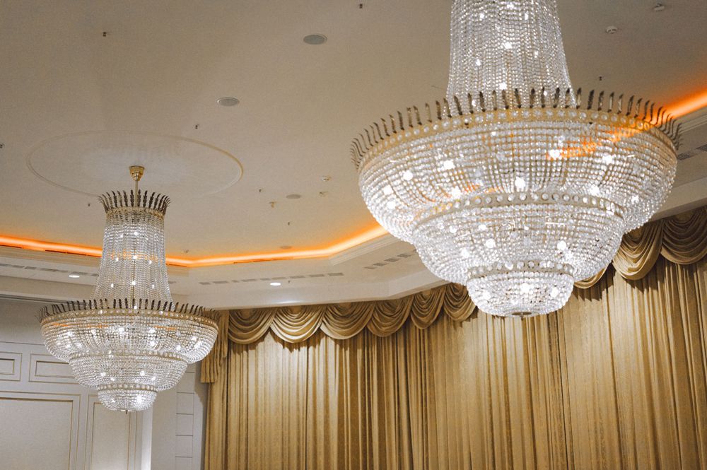 Elegant crystal chandeliers hang from an ornate ceiling in a luxurious ballroom with gold drapes.