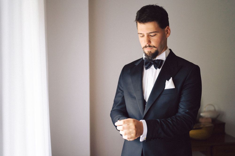 Person in black tuxedo and bow tie adjusting cufflinks by window.