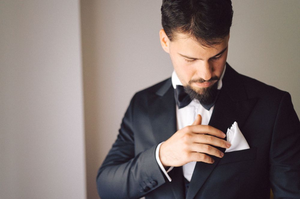 Groom in black tuxedo and bowtie adjusts his pocket square while getting ready for wedding ceremony.