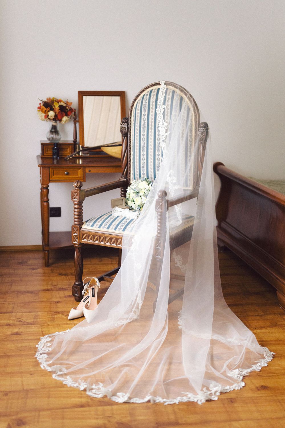 Wedding veil draped over vintage chair next to antique vanity with floral arrangement.